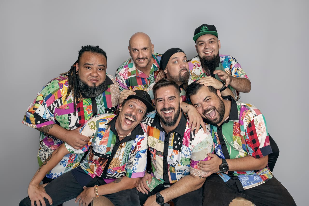 Os sete músicos da Confraria Musical abraçados e rindo, com as camisas de retalhos coloridos