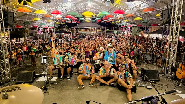 A banda sentada no palco, de costas para uma multidão de braços erguidos sob guarda-chuvas coloridos